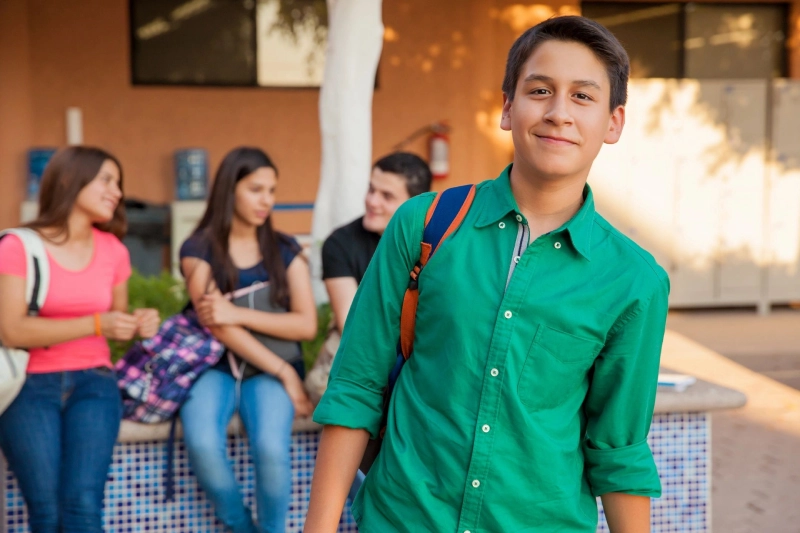 Smiling teenage boy with a backpack standing at a school campus, embodying the vibrant school culture, with peers in the background.