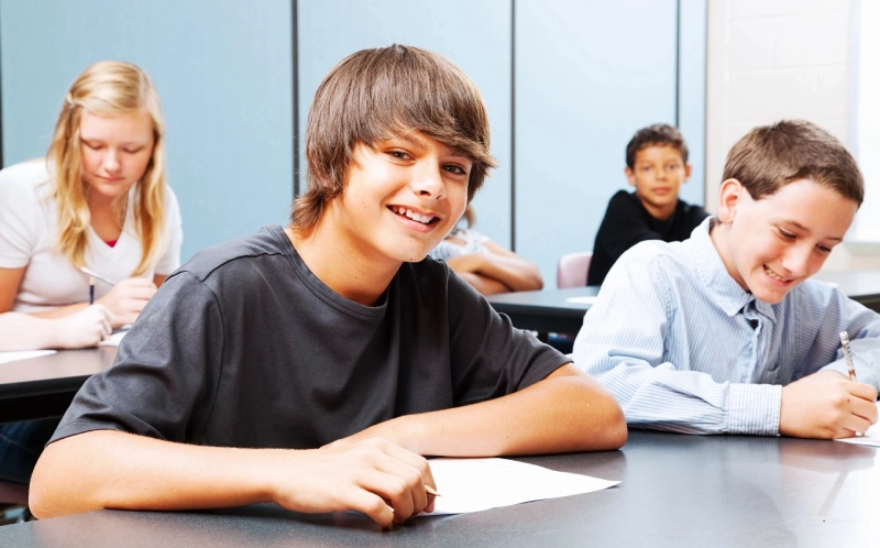 A smiling teenager looking at the camera while surrounded by other students who are writing.