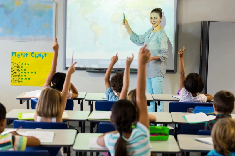 A teacher stands in front of a classroom, pointing to a map while discussing community involvement, as students raise their hands eagerly.